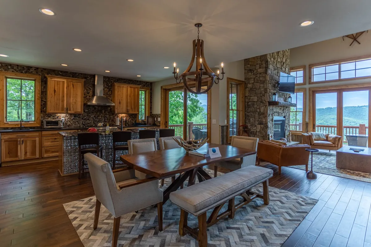 A spacious, open-concept dining area with a large table for six sits beneath a striking chandelier. The dining space flows into the living area, with a fireplace and wide windows offering sunset mountain vistas. The setting is ideal for family dinners, celebrations, and laughter-filled gatherings.