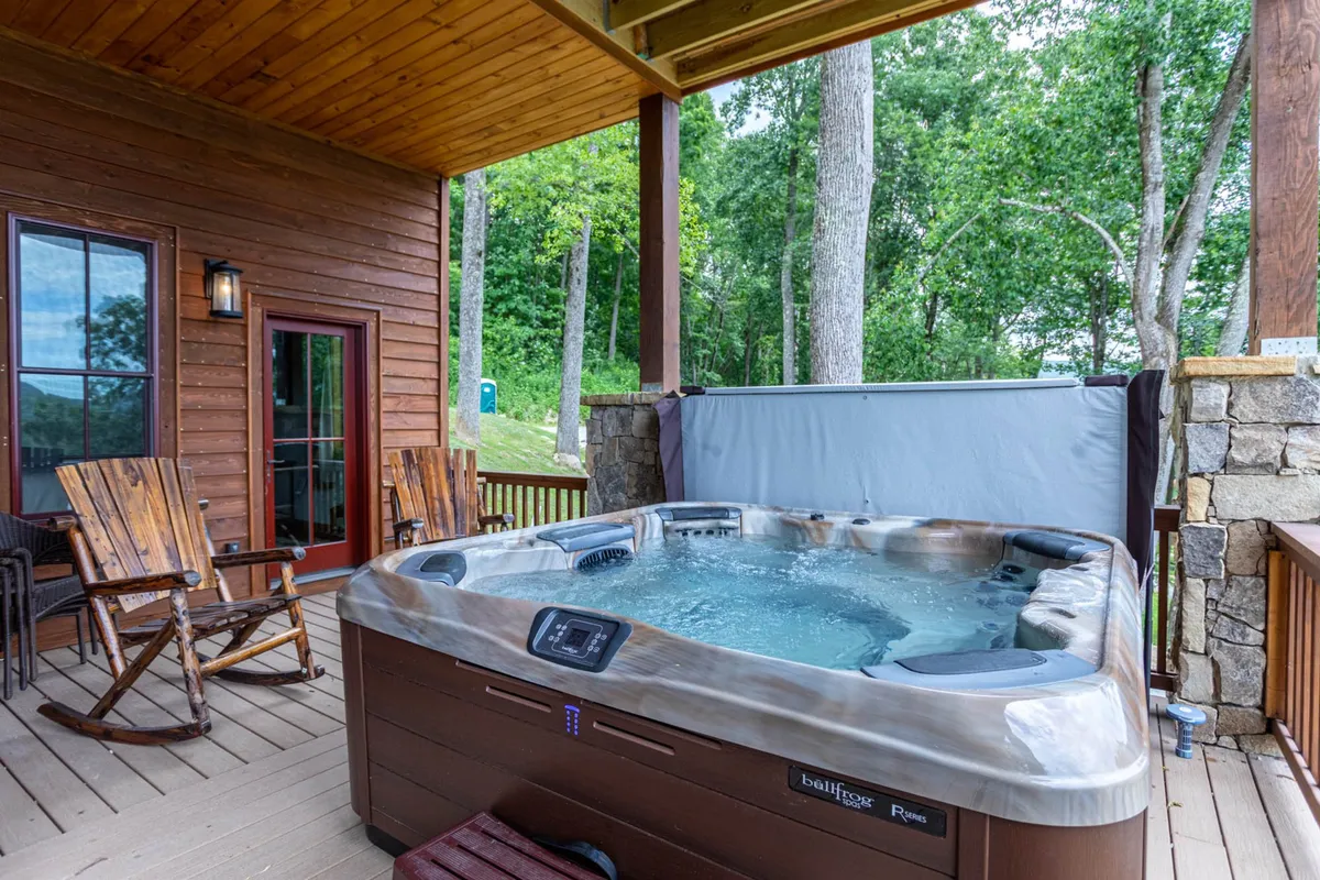 This exterior image features a wooden deck nestled amid natural greenery, with a hot tub and wooden rocking chairs positioned to enjoy the tranquil mountain setting. The inviting hot tub is the focal point, while the rocking chairs offer a cozy spot to enjoy coffee or evening chats, all surrounded by lush nature.