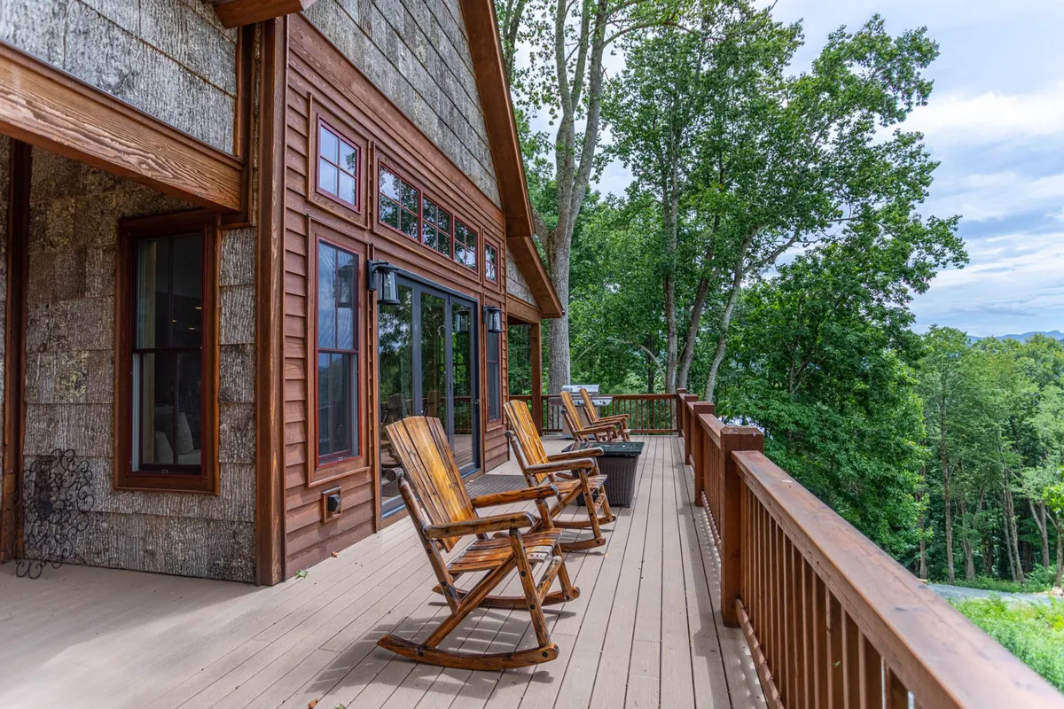A serene view of a wooden deck bordered by large windows and surrounded by trees. Wooden rocking chairs are set up to face the view, inviting couples to relax, socialize, or enjoy a peaceful moment together while immersed in nature.