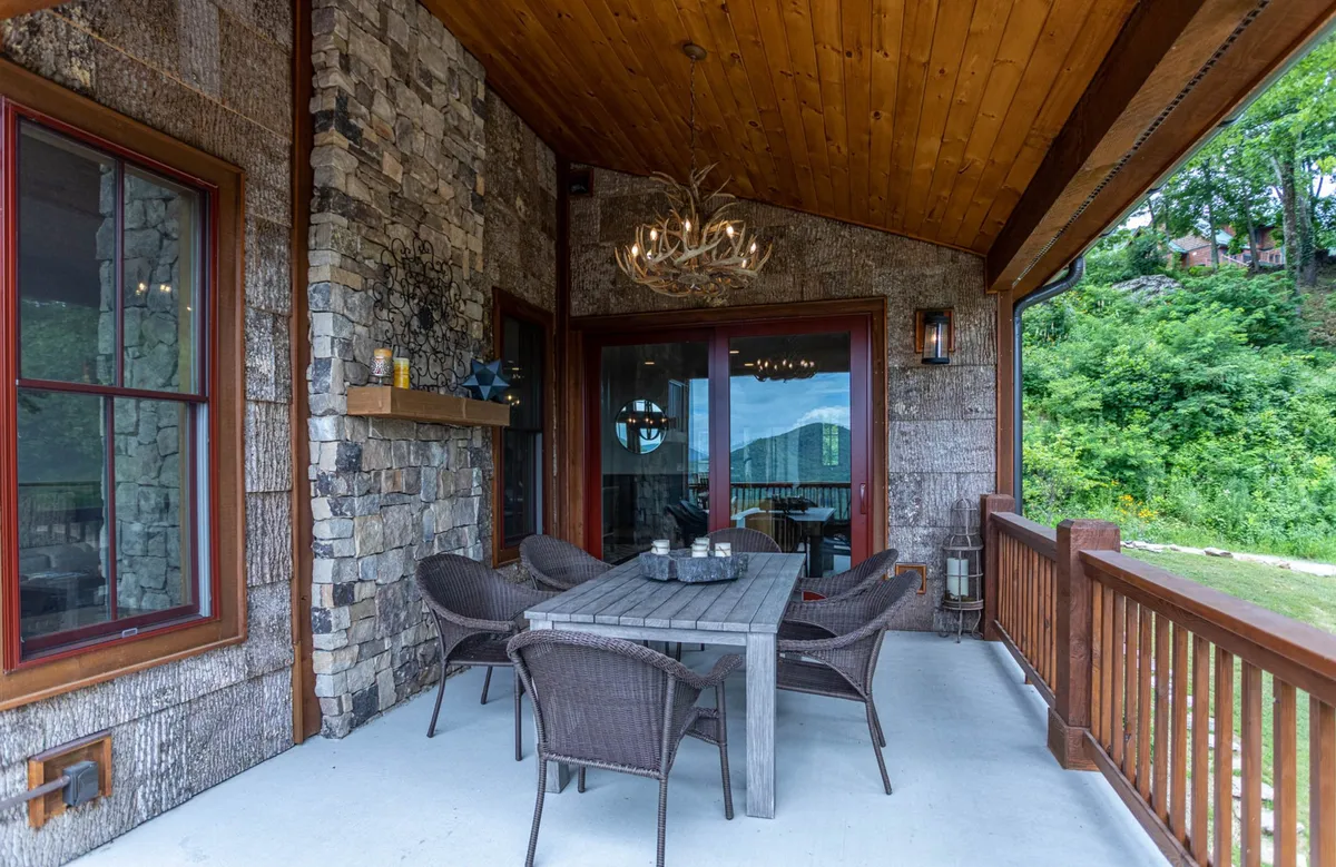 An inviting outdoor dining area with a stone wall, wooden ceiling, a large table, and elegant chandeliers. The scene looks through glass doors to the surrounding landscape, inviting group gatherings, celebratory dinners, and sociable evenings under the open sky.