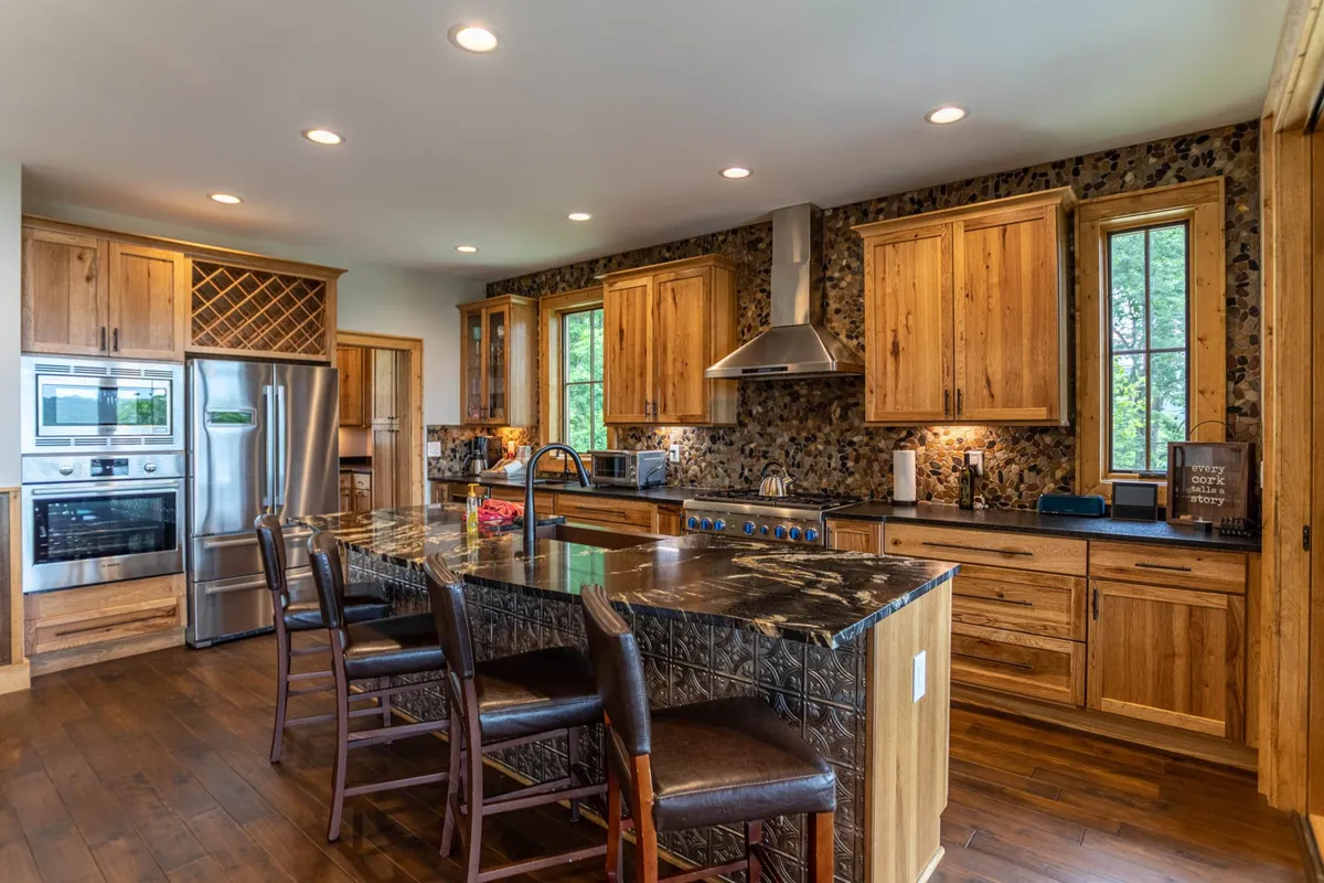 This image showcases the central island with seating, stainless steel appliances, wooden cabinetry, and a textured stone backsplash. The kitchen appears spacious and inviting, perfect for cooking and entertaining. The modern, upscale elements highlighted here directly reference the luxury appliances and open layout described in the section.