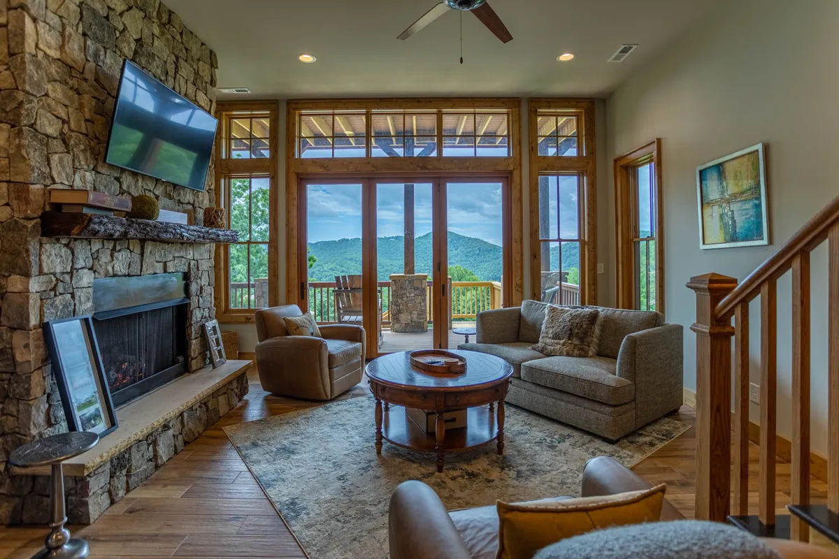 This interior shot highlights a warm, inviting living area with a stone fireplace, comfortable seating, and expansive windows framing breathtaking mountain views. The ambient lighting, plush furnishings, and visible television set the scene for relaxed movie nights or cozy conversations. Locally sourced artwork and wood accents evoke authentic Blue Ridge character.