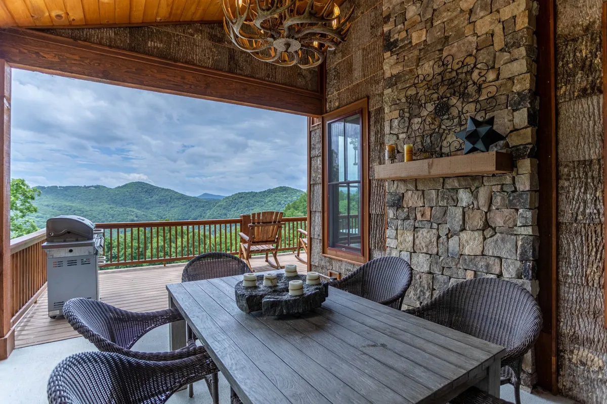 An outdoor dining setup on a covered deck features a sturdy wooden table with seating for six, a chandelier overhead, and panoramic views of the surrounding mountains. The inviting space hints at al fresco meals, barbecues, and relaxed mornings with coffee, all set against a backdrop of natural beauty.