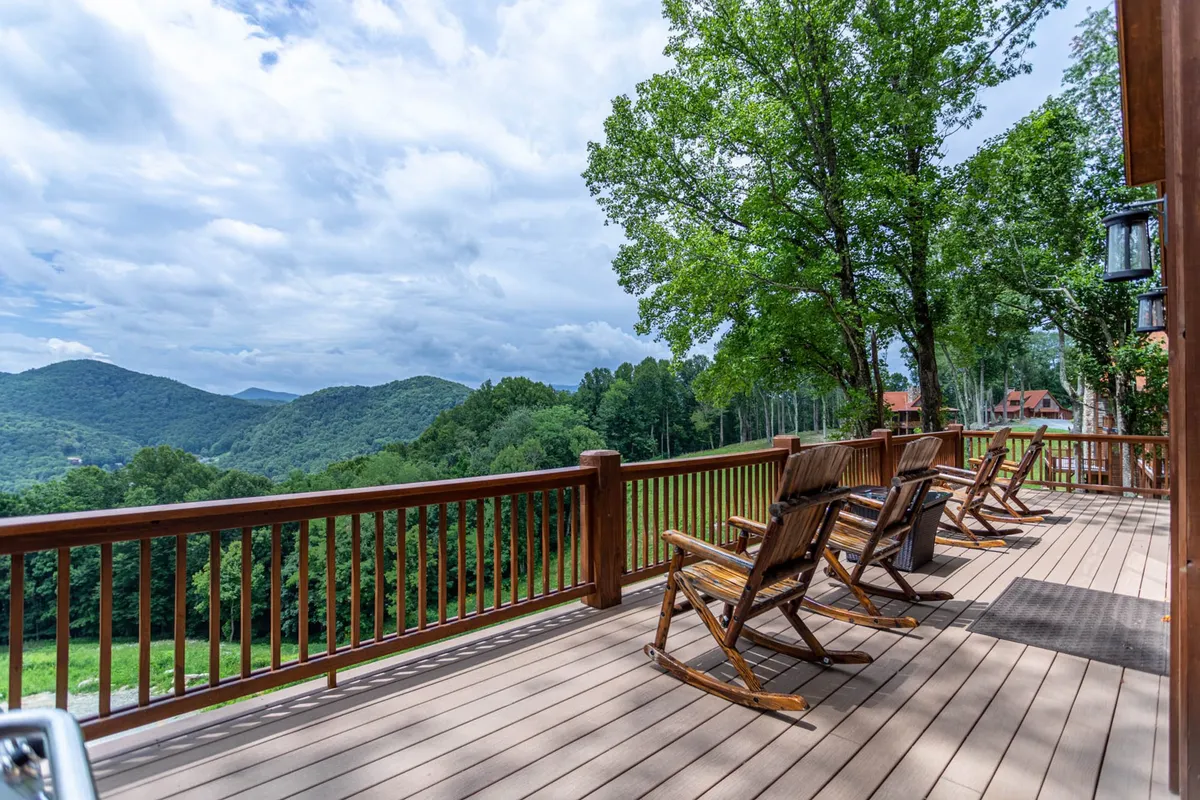 A peaceful exterior deck scene featuring wooden rocking chairs positioned to face a sweeping mountain view, surrounded by lush greenery and a classic deck railing. The early morning or late afternoon light hints at a tranquil outdoor experience, ideal for unwinding after an adventure or soaking in the mountain air.