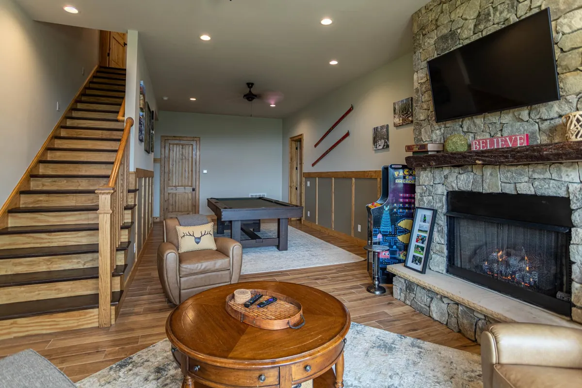 This image captures the vibrant lower-level game room featuring a pool table, a cozy fireplace, a large TV, and a classic arcade machine. The warm lighting and inviting furnishings create a welcoming environment for friendly competition and relaxation. The staircase and spacious layout hint at multi-generational gatherings and easy flow between spaces.