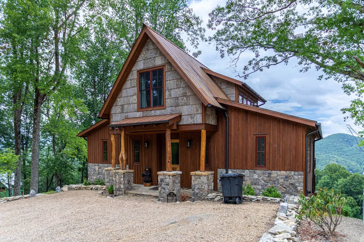 The image showcases the inviting exterior of Bear Tracks, a custom single-family house nestled among towering trees. The home's striking stone facade, rich wooden entrance, and expansive windows are set against a serene mountain backdrop. A spacious porch offers a perfect perch to take in the breathtaking mountain views, while the natural surroundings evoke tranquility and seclusion.