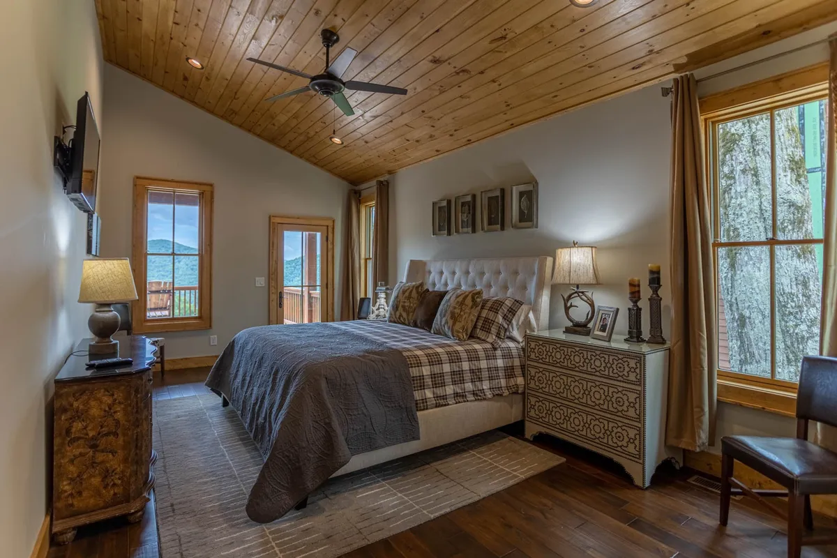 A tranquil king bedroom featuring a bed with patterned bedding, large windows that offer scenic views, a decorative lamp on the nightstand, and tasteful wall art. The space feels restful and private, bathed in natural light.