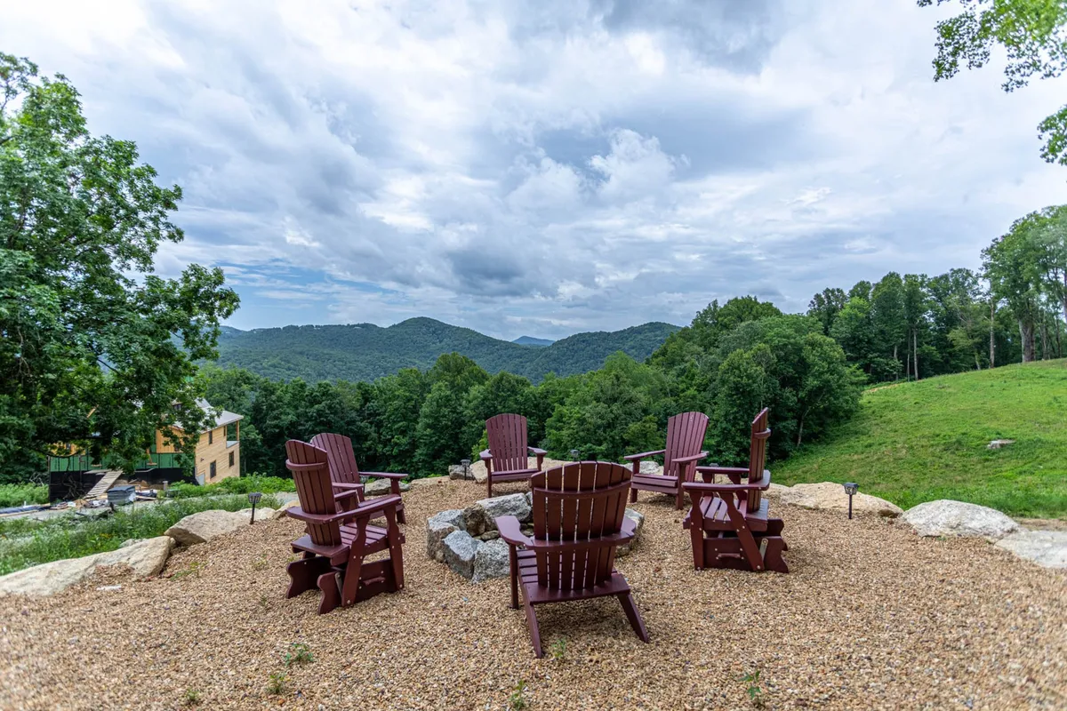 A serene fire pit area featuring wooden Adirondack chairs arranged around a central fire pit, framed by lush trees and mountain views. The space beckons for s’mores, stories, and communal relaxation.