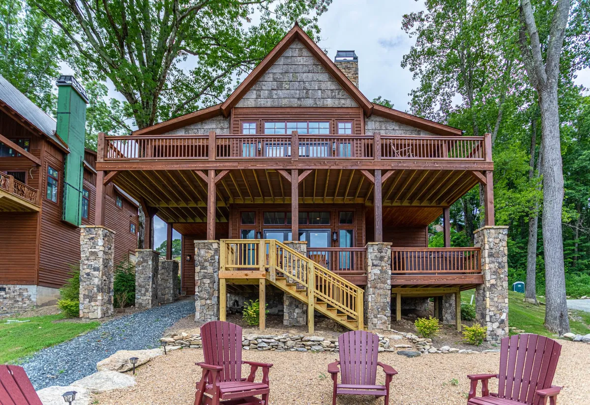 A spacious wooden deck with stone pillars and bright red Adirondack chairs overlooks a landscaped garden. The area is set up for outdoor gatherings, BBQs, and relaxing evenings around the fire pit, with ample room for families to socialize and share meals in a welcoming, communal space.